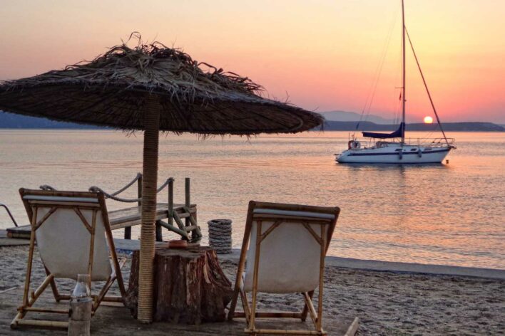 Wooden deck chairs under palm beach umbrella look out over yacht on water with a sunset sky