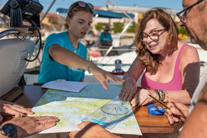 three people sit around a sailing chart. One person is pointing at something on the chart