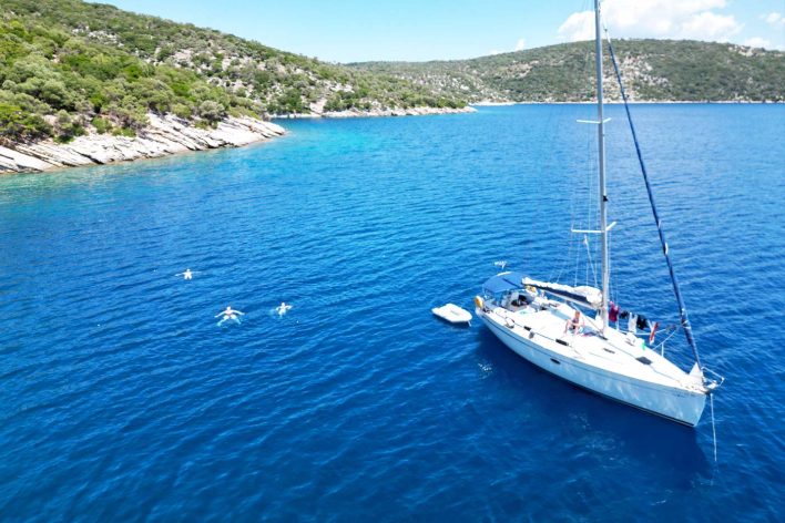 white sailing yacht moored at anchor in a bay. There are people swimming behind the yacht. The water is a deep blue colour. There is land in the background