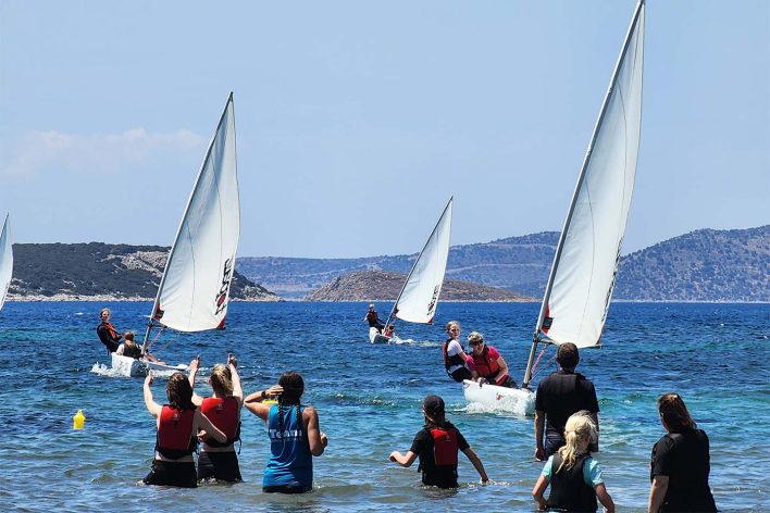 3x sailing dinghies sail towards a group of families stood in shallow water