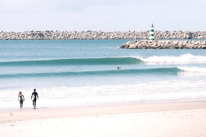 2 surfers walk into the shallows with waves at a beach club in Portugal